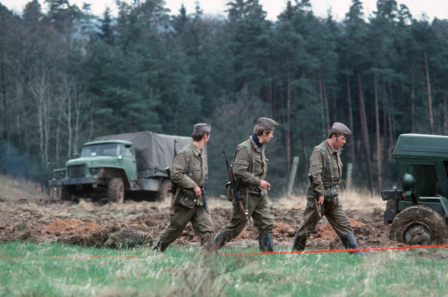 Three soldiers in Cold War uniform crossing a field past an army lorry